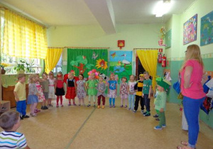 Dzieci w kolorowych ubrankach stoją ustawione w półkolu. Obok stoi nauczycielka.